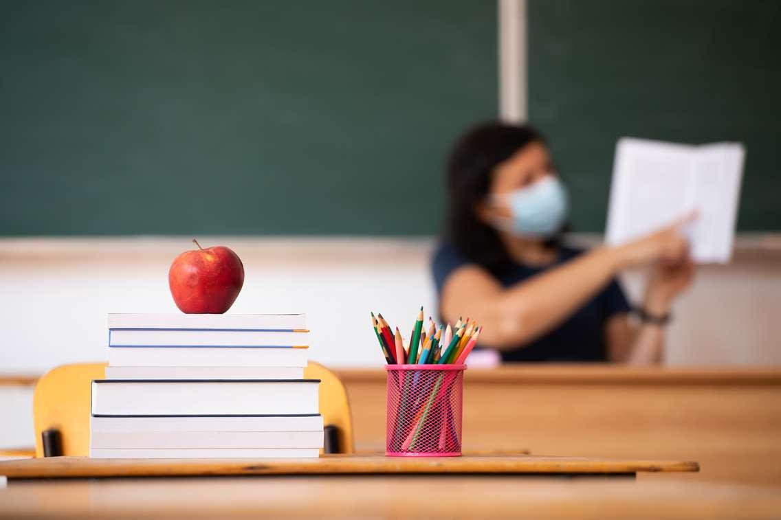 Back to school concept. apple, accessories and books in the classroom, teacher wearing mask in the background. © Can Stock Photo / erika8213