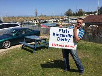 Travis Crawford, co-chair of the 2016 Kincardine Fishing Derby, holding the sign overlooking the Marina, and gesturing that it's the 31st annual event.